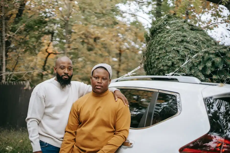 Bearded African American father embracing plump teenager near modern car with fir tree on roof while looking at camera