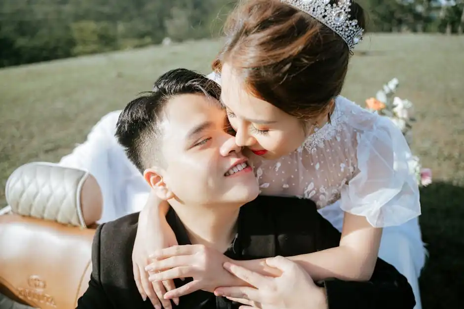 Happy Asian bride in white wedding dress and diadem sitting on back seat of retro car and embracing handsome young groom after wedding ceremony