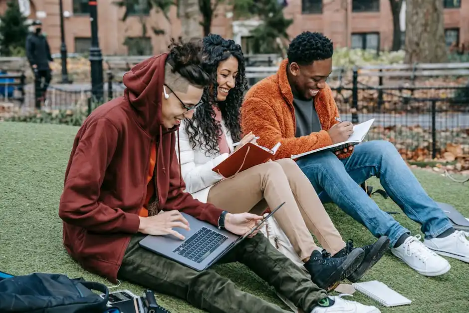 Happy multiracial group of students in casual clothes sitting together on grass and studying with laptop and books